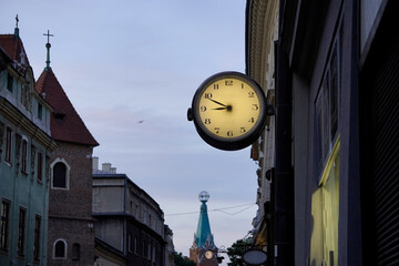 Vintage street clock glowing softly at dusk, historic buildings with detailed facades, pointed church tower with cross, soft evening sky, urban calmness, architectural blend.
