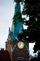 Clock tower with glowing face framed by dark green tree leaves, red brick facade, turquoise spire,...