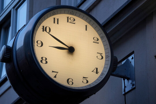 Large illuminated outdoor clock mounted on a building facade. The clock face is bright yellow with black numerals and hands, showing the time as approximately 9:50. The clock housing is black and atta