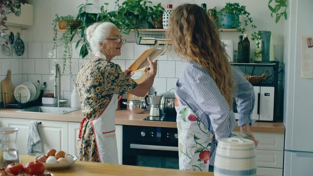 Young woman putting on apron in cozy home kitchen, smiling and reading old recipe book with loving grandmother while going to cook meal together