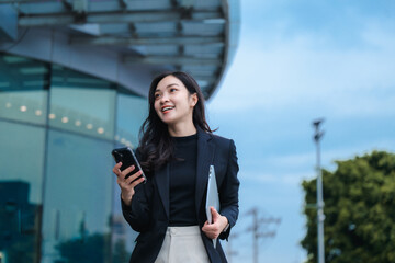 Smiling businesswoman using smartphone and holding laptop walking near office building