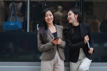 Two young businesswomen are using smartphone and laptop, talking and smiling near office building