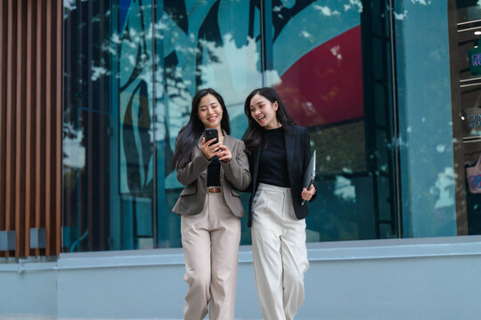 Two young businesswomen are walking and looking at smartphone in front of modern glass building