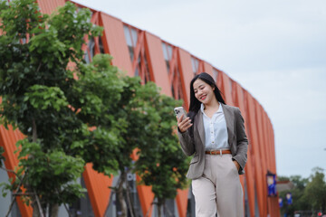 Smiling businesswoman using smartphone while walking near modern office building