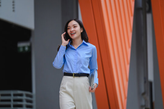 Businesswoman walking and talking on phone holding laptop in modern office district