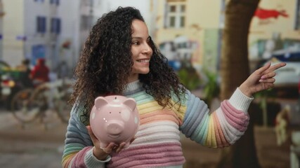 Young hispanic woman smiling on street holding piggybank wearing colorful sweater outdoors in urban area pointing happily. - Powered by Adobe
