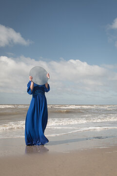 woman in long blue dress on the beach standing in shore holdint roud mirror hiding her face