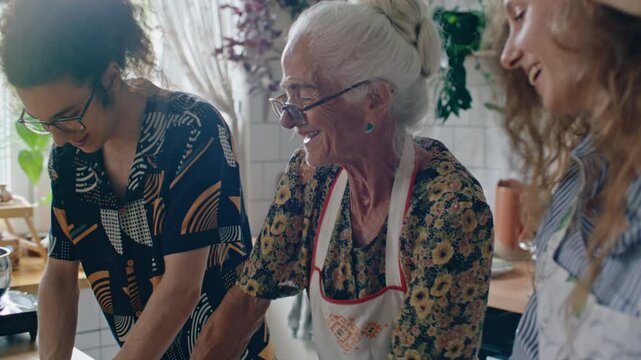 Elderly grandmother in apron smiling and talking to young grandkids as kneading dough together in home kitchen. Tilt-up shot
