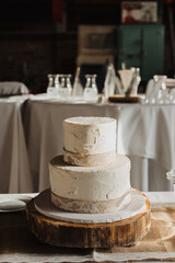 Rustic two-tier buttercream wedding cake with lace burlap ribbon on wooden stand in warmly lit reception setting with white table linens and glassware