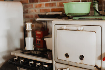Vintage kitchen interior featuring white enamel stove with worn handles, green mixing bowl on top, brick wall background, old cooking tools, and rustic nostalgic atmosphere