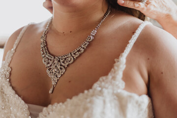 Close-up of ornate silver bridal necklace being fastened on bride wearing beaded white wedding dress in soft natural light capturing elegant and intimate preparation moment