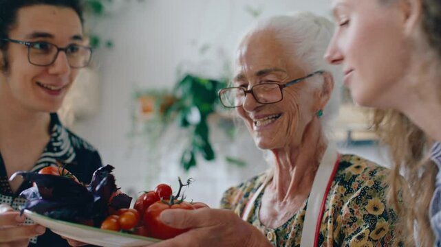 Joyful elderly woman holding bowl of fresh tomatoes and basil leaves while young grandchildren enjoying smell of fresh vegetables and herbs in sunlit kitchen
