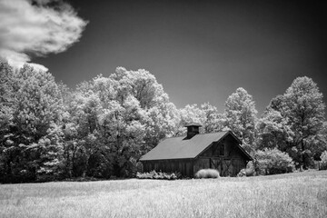 This striking black-and-white infrared photograph captures a rustic barn nestled in a serene countryside field, surrounded by lush, glowing trees under a dramatic sky , evoking timeless tranquility .