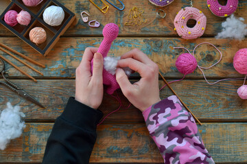Hands shaping neck of pink amigurumi dinosaur using crochet hook wool yarn and stuffing during handmade toy process on wooden table with accessories, Step 2 Stuff the toy with padding polyester