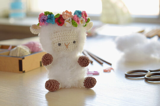 Handmade sheep amigurumi toy with colorful flower crown standing on desk near crochet tools and soft stuffing finished wool figure smiling close up crafting result - Powered by Adobe