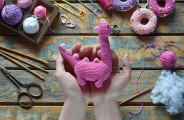 Amigurumi pink dinosaur toy shown in full held by hands above crafting table with yarn scissors stuffing and hook soft handmade crochet project completed