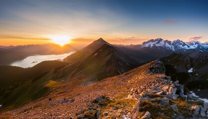 a dramatic mountain ridge leads toward a glowing sunset sky with lakes and distant snow capped peaks on the horizon