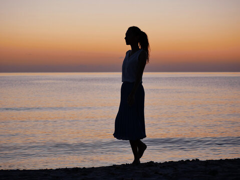 Silhouette of a woman walking along the beach at sunset and enjoying the peaceful sea view. Tranquil mood, calm water, colorful sky and relaxing summer scene