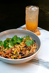 Asian beef udon noodles with vegetables, sesame, and fresh cilantro. Served with iced orange lemonade in a textured glass.