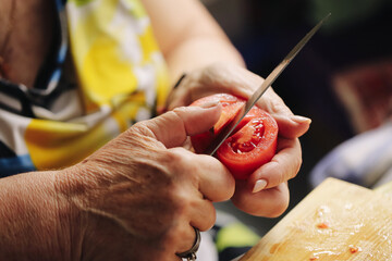 Senior woman slicing tomato. Close-up of hands preparing vegetables. Homemade cooking preparation....