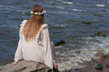 Young Woman in a White Dress and Daisy Flower Crown Sitting on a Boulder at the Rocky Shoreline of...