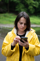 Vertical shot of woman in yellow raincoat scrolling phone in forest. Concept: digital connection in...