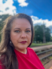 Confident woman with long dark hair, wearing a vibrant red shirt, poses outdoors against a bright...