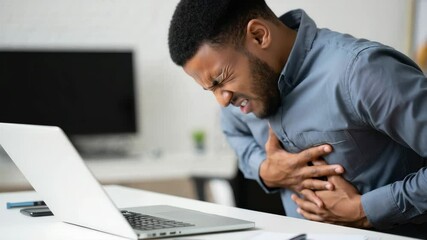 A man is seated at his desk, struggling with sudden chest pain as he works intently on his laptop. The office environment is modern, indicating a busy workday
