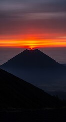 Dramatic sunrise view behind conical mountain silhouette with layers of misty ridges and glowing sky.