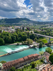 Naklejka premium Scenic view of a vibrant river flowing through lush green landscapes, with bridges connecting urban areas and dramatic clouds creating an atmospheric backdrop
