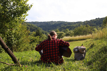 A young guy in a red shirt and jeans with a guitar travels alone through green mountains and nature.