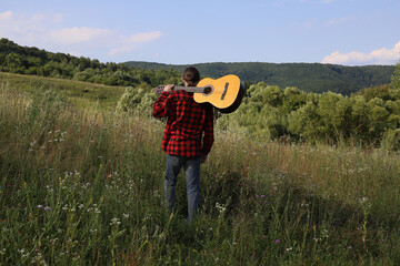 A young guy in a red shirt and jeans with a guitar travels alone through green mountains and nature.