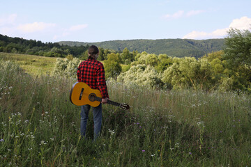 A young guy in a red shirt and jeans with a guitar travels alone through green mountains and nature.