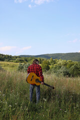 A young guy in a red shirt and jeans with a guitar travels alone through green mountains and nature.