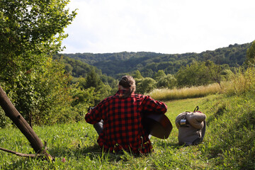 A young guy in a red shirt and jeans with a guitar travels alone through green mountains and nature.