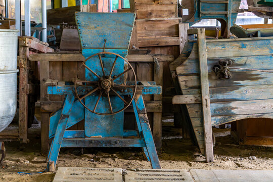 Old blue agricultural threshing machine with hand wheel among wooden crates in a rustic farm setting.