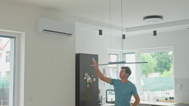 Man using remote control to operate wall-mounted air conditioner in modern kitchen. Person adjusting temperature for cooling in hot weather days