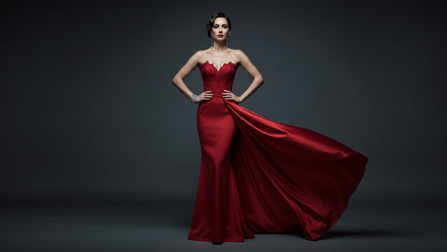 Elegant woman in luxurious red flowing evening gown posing confidently against a dark studio background, showcasing fashion and glamour.