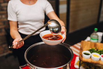 Photo of cook, waiter young woman pouring ukrainian fresh red borscht from big pot with spoon into plate in restaurant. Traditional dish concept.