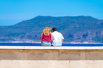 Unidentified young man and woman sitting and looking at blue sea water on Favignana island, near...
