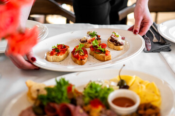 Close-up of a hand serving a white plate with assorted bruschetta topped with tomatoes, cheese, and greens on a rustic table, showcasing gourmet appetizer presentation.