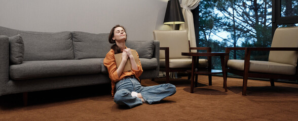 Young woman sitting on the floor in a cozy living room, wearing a stylish orange sweater and denim...