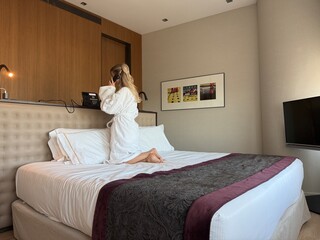 Woman in white hotel bathrobe talking on a landline phone while sitting on the bed. Possibly making...