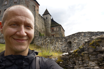 Selfie photo of a forty year old man with short hair exploring the sights of the Czech Republic. Selfie of a man with a stone castle behind his back. Traveler selfie while visiting a medieval castle.