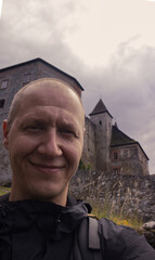 Selfie photo of a forty year old man with short hair exploring the sights of the Czech Republic. Selfie of a man with a stone castle behind his back. Traveler selfie while visiting a medieval castle.