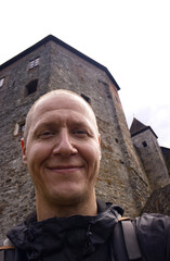 Selfie photo of a forty year old man with short hair exploring the sights of the Czech Republic. Selfie of a man with a stone castle behind his back. Traveler selfie while visiting a medieval castle.