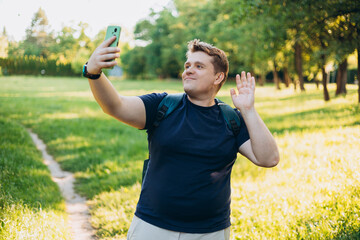 Young Caucasian blonde 30s man smiling confident making selfie at green park outdoors. Traveler. Summer time. Portrait of happy man in casual clothing standing on nature background