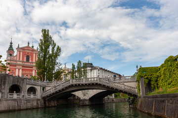 A classic view of Ljubljana, Slovenia, centered on the iconic Triple Bridge (Tromostovje) spanning the Ljubljanica River, under a partly cloudy sky