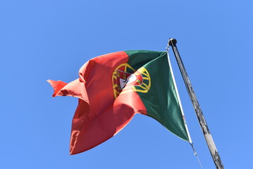 Drapeau du Portugal suspendu à un mât, sous un ciel bleu, suivant la direction du vent.
