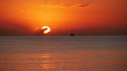 A breathtaking sunset over the calm sea with a sailboat on the horizon. Warm orange and golden tones fill the sky and reflect on the water. Travel, nature, relaxation, and maritime concept